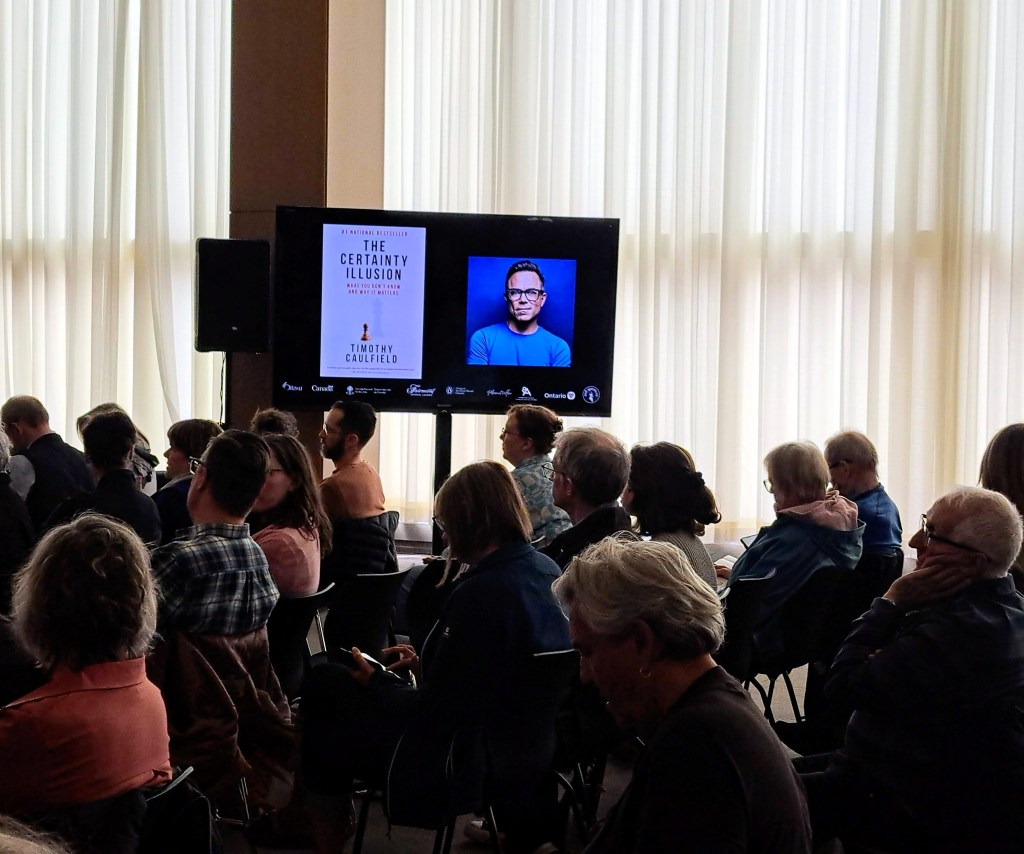 Colour photo of Timothy Caulfield projected onto a screen with audience present at Library and Archives Canada book event 